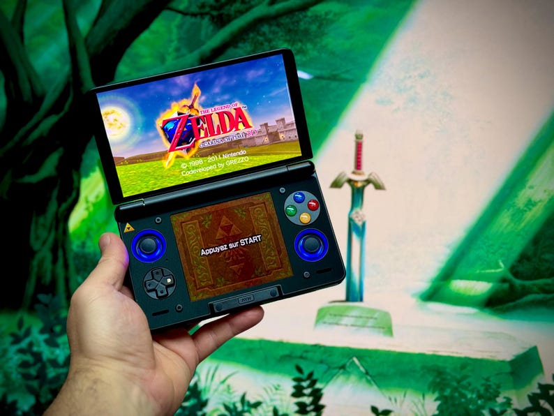 May include: A black Nintendo 3DS handheld game console showing a scene from "The Legend of Zelda". The top screen displays a castle and a logo. The bottom screen shows a game menu with the text "Appuyez sur START". A decorative sword is in the background.