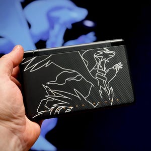 May include: Black handheld gaming console with a white line drawing of a dragon-like creature. The console is held in a hand against a dark background with a blue, blurred shape. The text "MON BLACK" is visible.
