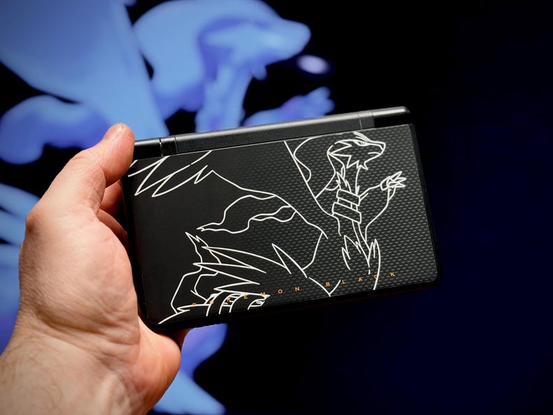 May include: Black handheld gaming console with a white line drawing of a dragon-like creature. The console is held in a hand against a dark background with a blue, blurred shape. The text "MON BLACK" is visible.