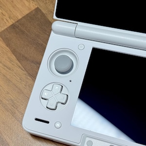 Pu&ograve; includere: Una console di gioco portatile bianca con un pad direzionale grigio, pulsanti e un joystick. La console ha uno schermo nero ed &egrave; parzialmente aperta. Il dispositivo &egrave; su una superficie di legno.