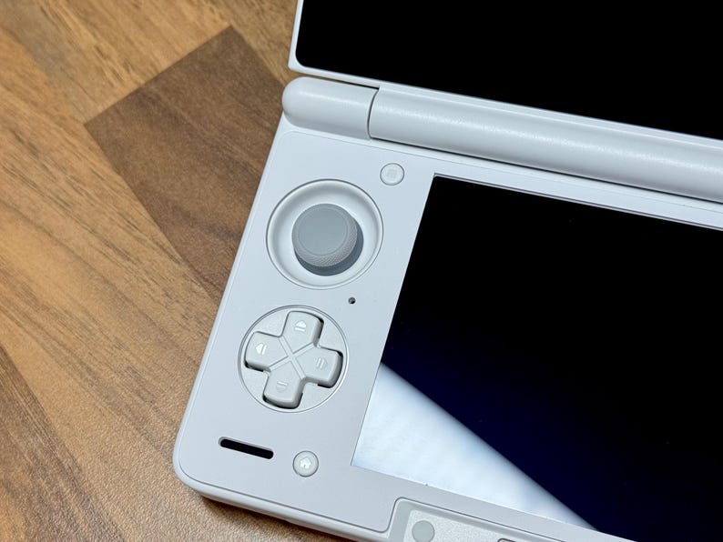 Pu&ograve; includere: Una console di gioco portatile bianca con un pad direzionale grigio, pulsanti e un joystick. La console ha uno schermo nero ed &egrave; parzialmente aperta. Il dispositivo &egrave; su una superficie di legno.