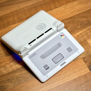 May include: A white handheld gaming console with a gray control panel. The console is open, revealing the buttons and the word "EJECT". The console is on a wooden surface.