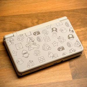 May include: A white Nintendo DS Lite handheld gaming console featuring a pattern of Mario characters and icons. The console is open, displaying the top screen and the patterned cover. The design includes Mario, mushrooms, stars, and other game elements.