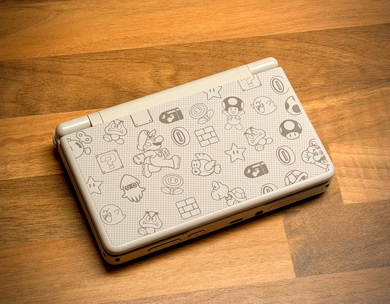 May include: A white Nintendo DS Lite handheld gaming console featuring a pattern of Mario characters and icons. The console is open, displaying the top screen and the patterned cover. The design includes Mario, mushrooms, stars, and other game elements.