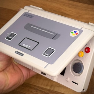 May include: A cream and grey handheld gaming device, styled after a Super Nintendo. The device has a screen that opens to reveal a directional pad, A and B buttons, and a joystick. The device has the Nintendo logo.