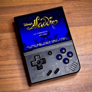 Op de afbeelding: Een zwart-grijze handheld gameconsole met het Disney's Aladdin-spel op een blauw scherm. De console heeft een D-pad, knoppen en de tekst "Commencer" en "Options".