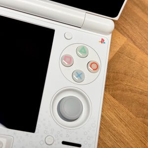 Op de afbeelding: Witte draagbare gameconsole met een zwart scherm en kleurrijke knoppen. De console heeft een D-pad, actieknoppen en een joystick. Het PlayStation-logo is zichtbaar. De console staat op een houten oppervlak.