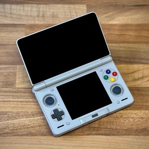 Peut inclure: Console de jeu portable gris clair avec deux écrans noirs, un ouvert et un fermé. La console a un pavé directionnel, des boutons colorés et deux joysticks. L'appareil est posé sur une surface en bois.