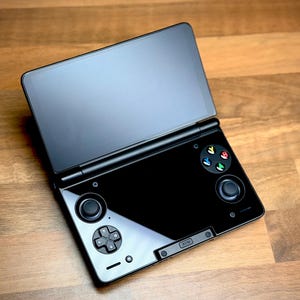 Pode incluir: Console de jogos portátil preta com design articulado e dois ecrãs. O ecrã inferior apresenta um painel direcional e botões de controlo. O ecrã superior está aberto, exibindo uma superfície escura. A consola repousa sobre uma superfície de madeira.