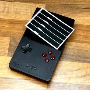 Peut inclure: Une console de jeu portable noire avec des boutons rouges et une feuille d'autocollants blanche avec des rayures noires sur le dessus. La console porte l'inscription "Analogue Pocket" sur le devant.