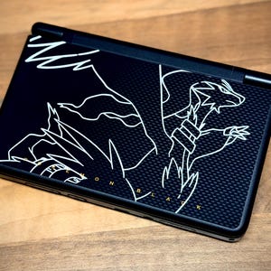 May include: Black handheld gaming console featuring a white outline illustration of a dragon. The console has a textured surface and the text "Pokemon Black" in gold. The device is on a wooden surface.