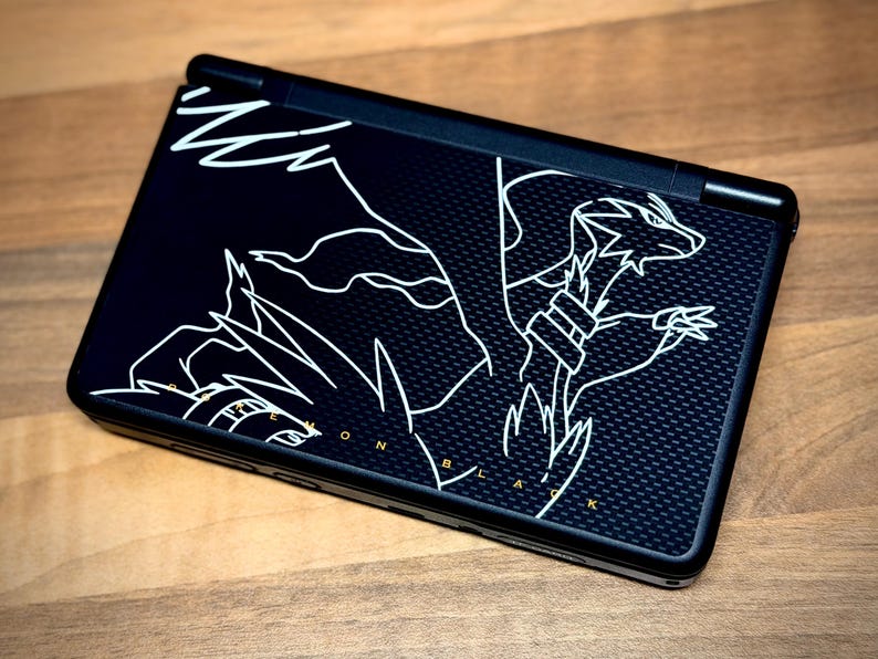 May include: Black handheld gaming console featuring a white outline illustration of a dragon. The console has a textured surface and the text "Pokemon Black" in gold. The device is on a wooden surface.