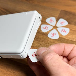 May include: A white handheld gaming console with a smooth finish, positioned next to five white guitar picks arranged in a flower pattern. Each pick features a small red cherry graphic. One additional matching cherry pick is held near the console.