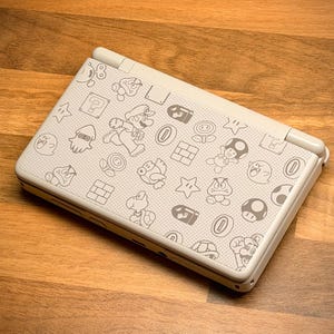 May include: A white handheld gaming console featuring a pattern of gray cartoon characters and icons. The design includes images of Mario, mushrooms, stars, and other elements from the Super Mario Bros. video game series. The console is on a wooden surface.