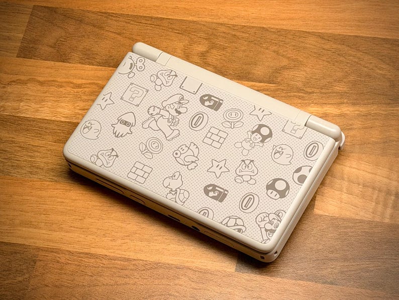 May include: A white handheld gaming console featuring a pattern of gray cartoon characters and icons. The design includes images of Mario, mushrooms, stars, and other elements from the Super Mario Bros. video game series. The console is on a wooden surface.