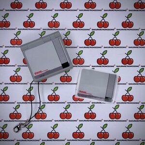 Puede incluir: Una consola port&aacute;til Nintendo Entertainment System gris y blanca con una correa negra. La consola est&aacute; sobre un fondo blanco con un patr&oacute;n repetitivo de cerezas rojas. La consola tiene una pegatina que dice "Nintendo Entertainment System".