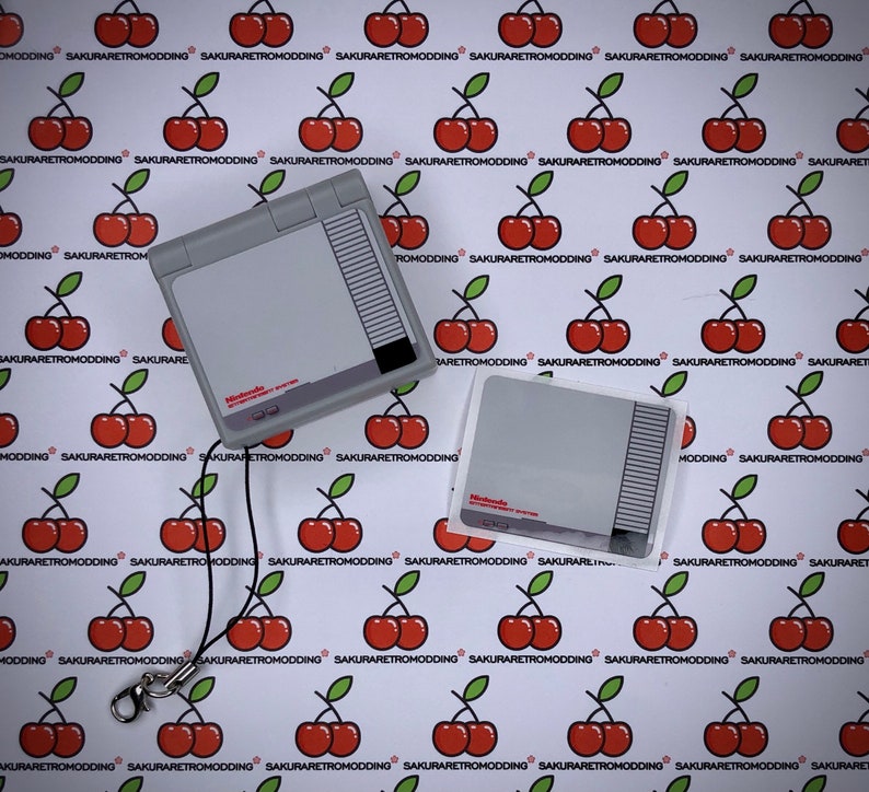 Puede incluir: Una consola port&aacute;til Nintendo Entertainment System gris y blanca con una correa negra. La consola est&aacute; sobre un fondo blanco con un patr&oacute;n repetitivo de cerezas rojas. La consola tiene una pegatina que dice "Nintendo Entertainment System".