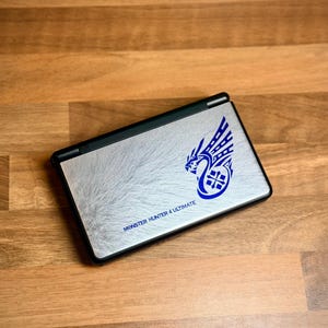 May include: A silver Nintendo DS Lite console featuring a blue dragon graphic and the text "MONSTER HUNTER 4 ULTIMATE". The closed console sits on a wooden surface.