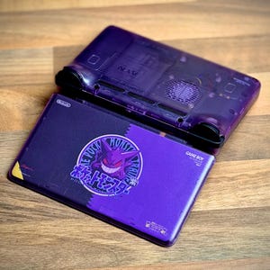 May include: A purple Game Boy Advance SP console with a custom design featuring a Gengar graphic and Japanese text. The console is open, revealing the internal components of the top half. The design is on a wooden surface.