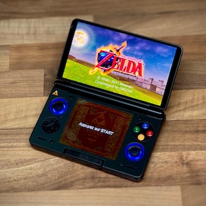 May include: A black handheld games console showing the game "The Legend of Zelda: Ocarina of Time 3D". The console has blue illuminated joysticks and buttons. The screen displays a colourful landscape with text in French: "Appuyez sur START".