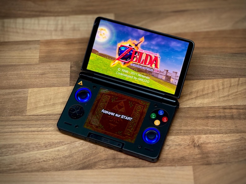 May include: A black handheld games console showing the game "The Legend of Zelda: Ocarina of Time 3D". The console has blue illuminated joysticks and buttons. The screen displays a colourful landscape with text in French: "Appuyez sur START".