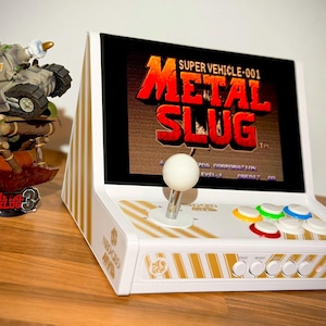 Op de afbeelding: Witte arcade console met "METAL SLUG" op het scherm. Voorzien van joystick, gekleurde knoppen en een tankbeeldje. Het retro design is versierd met gouden strepen.