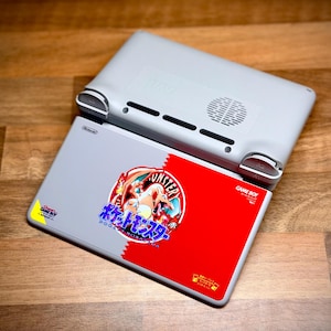 May include: A light gray Nintendo Game Boy handheld console with a red and gray cartridge featuring Japanese text and a cartoon graphic. The console is resting on a wooden surface.