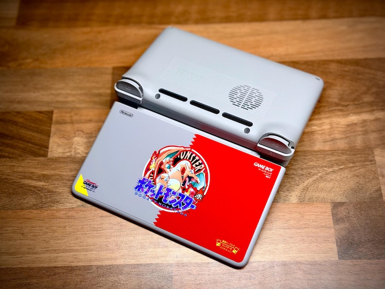 May include: A light gray Nintendo Game Boy handheld console with a red and gray cartridge featuring Japanese text and a cartoon graphic. The console is resting on a wooden surface.