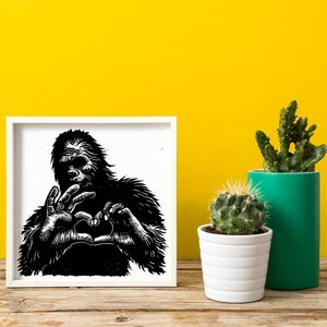 Può includere: Illustrazione in bianco e nero di un Bigfoot che fa un cuore con le mani. L'illustrazione è incorniciata in una cornice bianca e si trova su una superficie di legno.