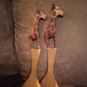 May include: A set of two wooden salad servers shaped like giraffes. The servers are brown and have a giraffe pattern. The server on the left is a spoon and the server on the right is a fork.