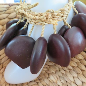 May include: A necklace featuring dark brown, teardrop-shaped beads suspended from a woven, beige rope. The beads are smooth and glossy, and the rope is knotted at the top. The necklace is displayed on a white form, with a woven background.