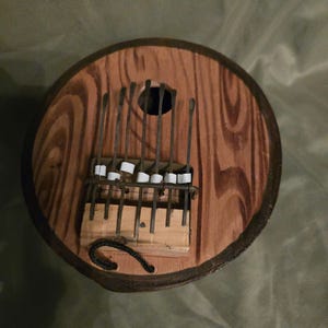 Peut inclure: Un kalimba en bois avec un corps rond en bois brun et une cordelette noire attachée. Le kalimba a une rangée de lames métalliques avec des élastiques blancs sur chaque lame.