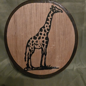 Peut inclure: Une plaque ronde en bois avec une silhouette noire de girafe sur un fond brun clair. La plaque a une bordure brun foncé.
