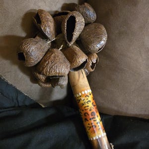 May include: A percussion instrument featuring a cluster of brown, textured, hollow seed pods attached to a cylindrical wooden handle. The handle displays a yellow and orange leopard print pattern with white dot accents and a woven rope base.