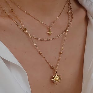 Può includere: Un set di collane dorate a strati composto da tre catene distinte. La catena più corta ha un piccolo ciondolo a forma di stella, la catena centrale è a maglie tipo graffetta e la più lunga presenta piccole perline e un ciondolo a forma di sole.