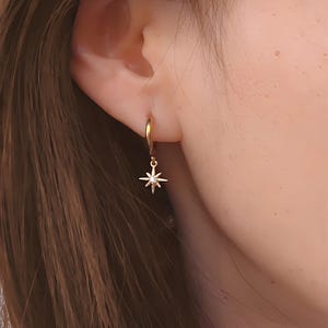 Peut inclure: Boucles d'oreilles créoles dorées avec un pendentif en forme d'étoile orné d'une petite pierre transparente au centre.