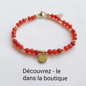 K&ouml;nnte beinhalten: Ein rotes Perlenarmband mit goldfarbenen Akzenten und einem kleinen, runden Goldanh&auml;nger. Das Armband besteht aus kleinen, runden roten Perlen und goldfarbenen Metallperlen. Der Text "D&eacute;couvrez - le dans la boutique" ist sichtbar.