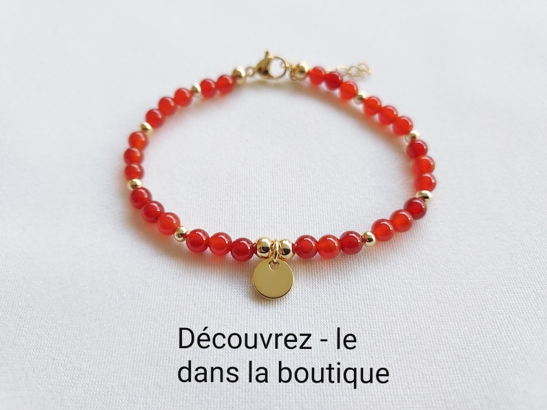 K&ouml;nnte beinhalten: Ein rotes Perlenarmband mit goldfarbenen Akzenten und einem kleinen, runden Goldanh&auml;nger. Das Armband besteht aus kleinen, runden roten Perlen und goldfarbenen Metallperlen. Der Text "D&eacute;couvrez - le dans la boutique" ist sichtbar.
