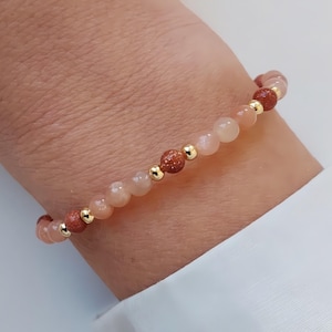 Peut inclure: Un bracelet délicat composé de perles couleur pêche et de pierres dorées, entrecoupées de petites perles dorées. Le bracelet est monté sur un élastique et est présenté sur un poignet.