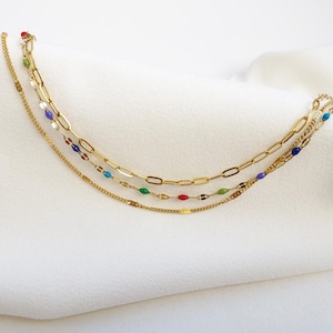 May include: A layered gold-toned necklace with three strands. The top strand features large oval links, the middle strand has small colorful beads, and the bottom strand is a delicate chain. The necklace has red, green, blue, and purple accents.