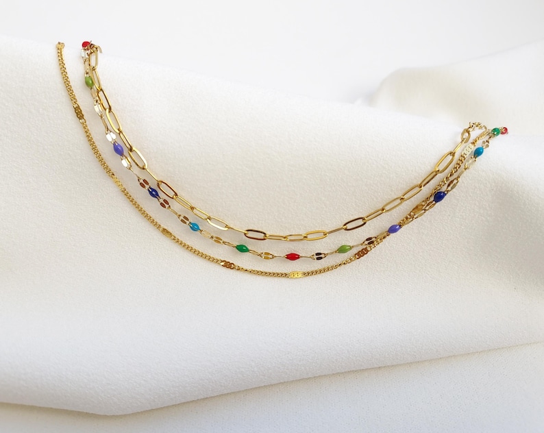 May include: A layered gold-toned necklace with three strands. The top strand features large oval links, the middle strand has small colorful beads, and the bottom strand is a delicate chain. The necklace has red, green, blue, and purple accents.