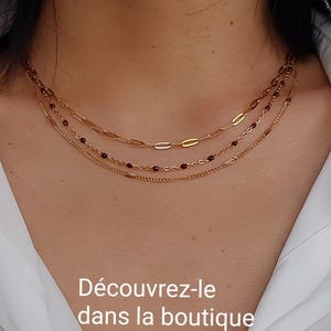 May include: Three gold necklaces layered on a person's neck. The top necklace has oval links, the middle one has small red beads, and the bottom one has a beaded design. The text "D&eacute;couvrez-le dans la boutique" is at the bottom.