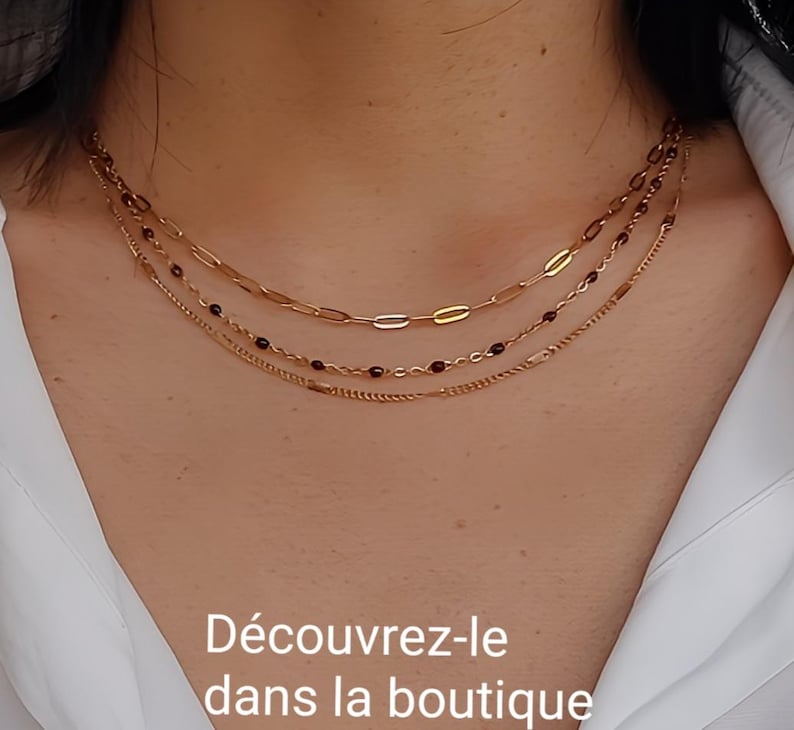 May include: Three gold necklaces layered on a person's neck. The top necklace has oval links, the middle one has small red beads, and the bottom one has a beaded design. The text "D&eacute;couvrez-le dans la boutique" is at the bottom.