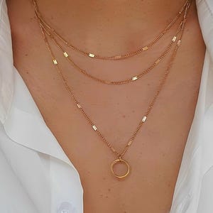 Peut inclure: Un collier doré à plusieurs rangs, composé de trois chaînes délicates. Les deux chaînes supérieures sont plus courtes et présentent de petits accents rectangulaires. La chaîne inférieure comporte un petit pendentif rond. Le collier est porté sur une chemise blanche.