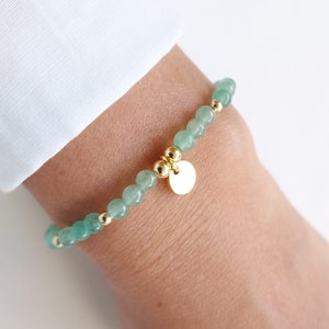 K&ouml;nnte beinhalten: Ein Armband mit hellgr&uuml;nen, runden Edelsteinperlen und goldfarbenen Akzenten. Das Armband hat kleine goldene Perlen und einen goldenen Anh&auml;nger. Das Armband wird am Handgelenk getragen.
