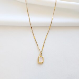 May include: A gold-colored necklace with a rectangular pendant. The pendant has a light pink center and is framed by a gold border. The chain is delicate and features small rectangular accents.