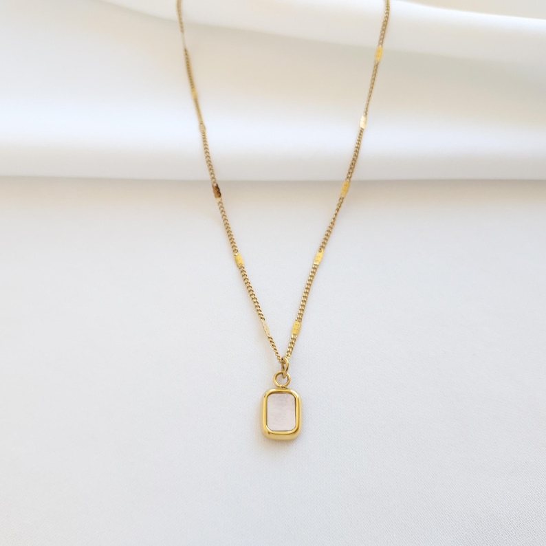May include: A gold-colored necklace with a rectangular pendant. The pendant has a light pink center and is framed by a gold border. The chain is delicate and features small rectangular accents.