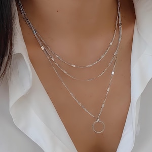 Può includere: Una collana a strati color argento composta da tre catenine delicate di diverse lunghezze. Le catene sono decorate con piccoli inserti metallici rettangolari e piatti, distanziati uniformemente, e la catena più lunga presenta un ciondolo circolare.