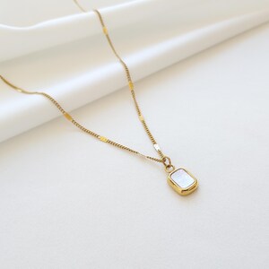 May include: A gold-colored necklace with a rectangular pendant. The pendant has a white, iridescent center and a gold-colored frame. The chain is delicate and has a subtle, textured design. The necklace is displayed on a white background.