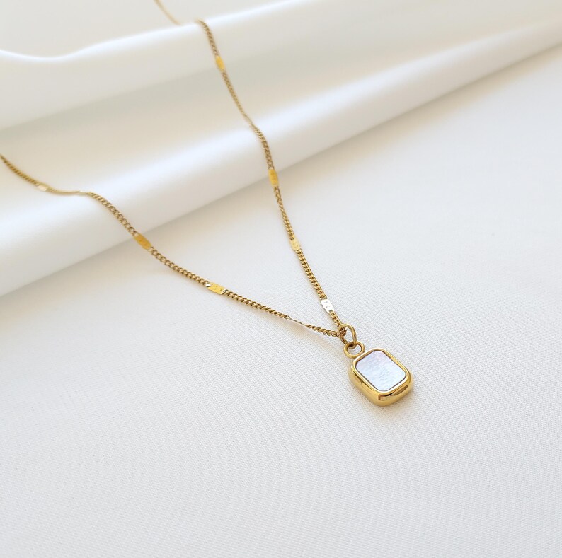 May include: A gold-colored necklace with a rectangular pendant. The pendant has a white, iridescent center and a gold-colored frame. The chain is delicate and has a subtle, textured design. The necklace is displayed on a white background.